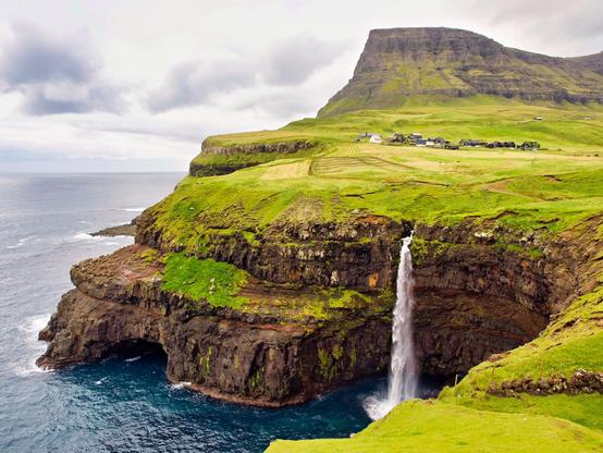 Rugged coastline of dark reddish cliffs falling into a dark blue ocean. The landscape is green by grass, in the background a flat top mountain stands up under an overcasted sky. On the right a waterfall is pouring directly into the ocean. The scene is serene and dramatic at the same time.