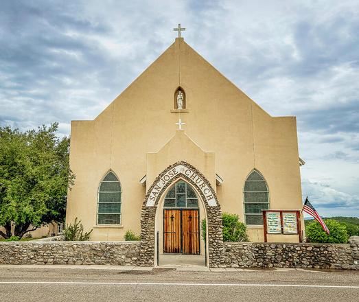 A historic church building stands with a beige facade and arched windows, featuring a small statue in a niche above the entrance. The church is named "San Jose Church" and includes a wooden gate and a stone wall in front. Photography by Debra Martz
