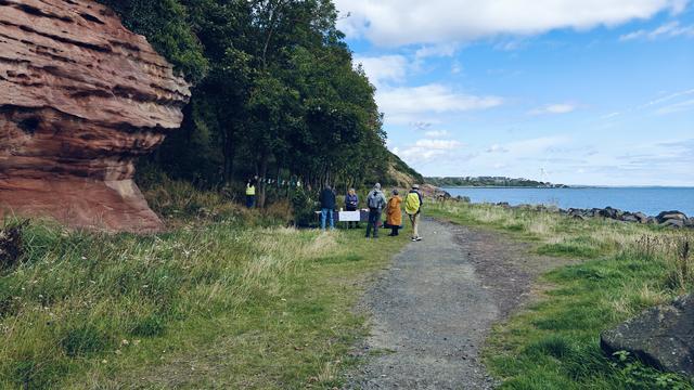 A path, with the sea to the right and red sandstone cliffs to the left. In the distance is a group of people gathered in front of a table