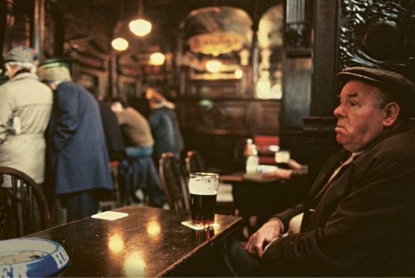 Foto di Douglas Corrance, The Pub, Glasgow, 1970s. L’immagine mostra una scena interna, probabilmente in un pub o bar. In primo piano c'è una persona seduta a un tavolo del pub, dove ci sono una pinta di birra scura e un posacenere. Sullo sfondo, altri clienti sono in piedi al bancone, e una persona indossa un cappello. L’ambiente ha un' atmosfera vintage o classica, suggerita dallo stile degli abiti e dal design degli interni, che include pannelli di legno scuro e dettagli ornati.