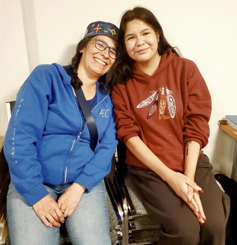 Two Indigenous women cousins, sitting side by side on stacked chairs. Elaine, on the left, is wearing a traditional Indigenous Anishinaabe hat, blue zip hoodie & jeans. Kristina, on the right, is wearing a burgundy hoodie with Indigenous feathers art design & black jogging pants. Both are smiling at the camera; Elaine is leaning her head on Kristina's shoulder.