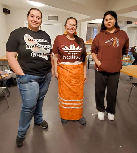 3 Indigenous women standing in workshop room.
Arianna(on the left), in jeans & t-shirt that says: Beautiful Indigenous Creature.
Kecia (in middle), wearing her newly made orange & white ribbon skirt with burgundy t-shirt with feathers spread like the Adidas logo with native in text below.
Kristina (on the right), wearing burgundy hoodie with Indigenous feathers design & black jogging pants.