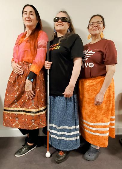 3 Indigenous women wearing their own newly made ribbon skirts. They're standing sideways, together, against a white wall.