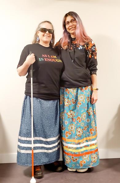 Two Indigenous women standing against white wall, wearing their own, newly made, ribbon skirts.