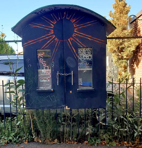 Black little free library, with arched roof, mounted on a black iron gate, in front of a parking lot. It has chain link to open/close.