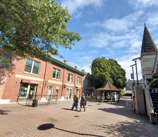 View of Fernwood Square. Old brick building on the right. Public gazebo in middle of square. Restaurant patio beside gazebo. To the left is the Belfry Theatre building. A couple is walking through middle of the square.
Sunny day.