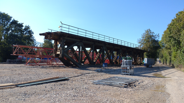 Ein Stahlfachwerkbrückenträger steht auf einer Baustelle rum. Davor ein Kasten mit länglichen Gasflaschen, dahinter Teile eines zerlegten Fachwerk-Kranenmastes.