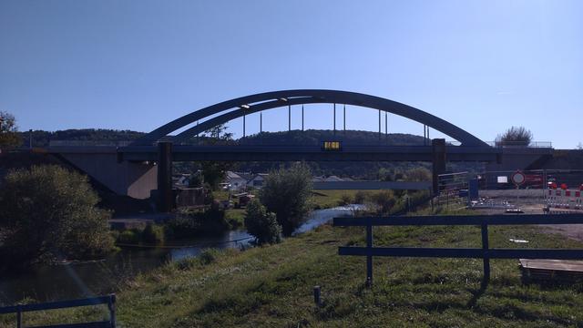 Eine Stahlbogenbrücke auf Brückenköpfen aus Beton über einen begradigtes Flüsschen. Die Fahrbahn ist an senkrechten Stahlseilen am Stahlbogen aufgehängt.