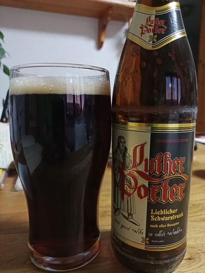 Auf einem Tisch steht ein Glas mit fast schwarzem Bier gefüllt und da eben die Bierflasche