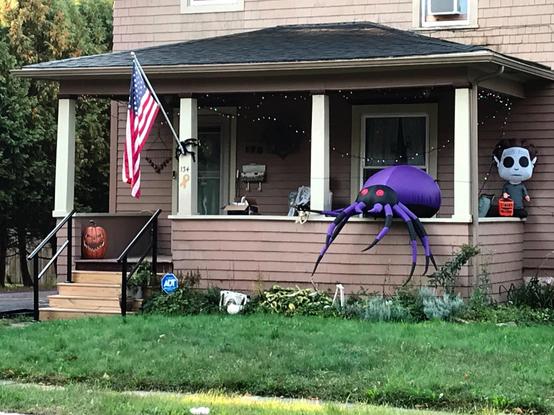 The front porch of a beige house. In addition to an American flag, the porch is festively adorned with Halloween decorations, including a giant black and purple spider with red eyes, some kind of pale undead looking fellow going trick or treating, and a rather malevolent jack-o'-lantern by the steps. There are also multicolored holiday lights strung about.