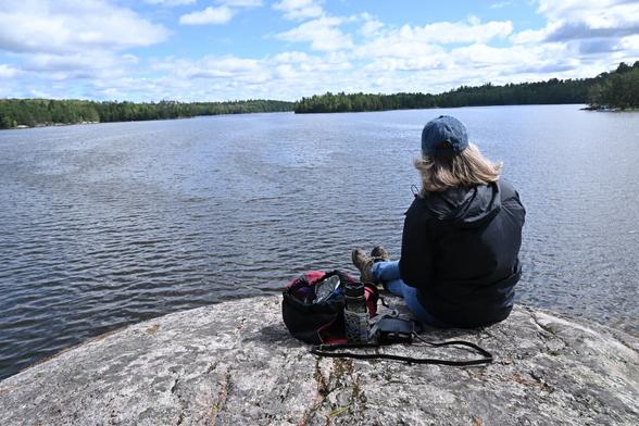 A woman sitting on a granite rock looking out across a northern lake. The sky has fluffy clouds.