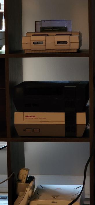 A shelving unit with multiple game consoles. On top is a super nintendo with a game that contains a translucent shell. On the middle shelf are two original Nintendos, the top one with a very black paint job, and a regular one below that. At the bottom is a sega dreamcast with a controller