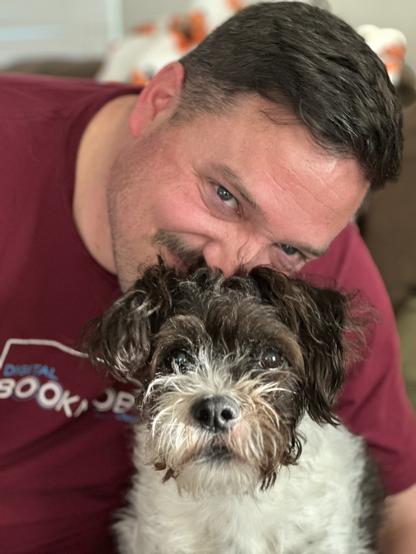 A man with short hair and a maroon shirt is playfully leaning towards a small, scruffy dog, which has a black and white coat. The background shows a cozy indoor setting.