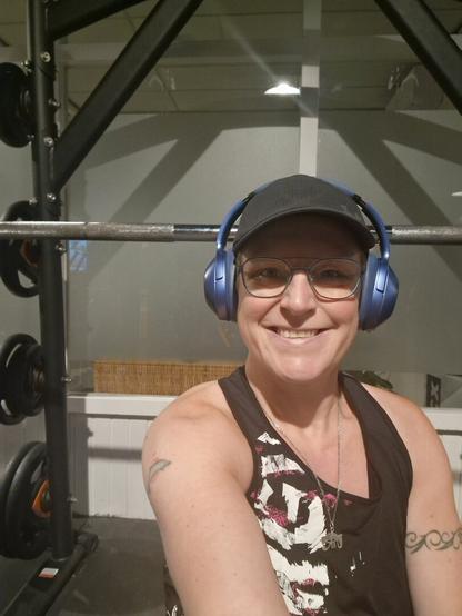 A selfie taken from the incline bench at the Smith Machine in the gym. Smiling at the camera. Wearing a black top with white accents, black cap, blue headphones and blue-rimmed glasses.