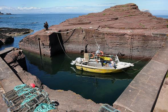 Seacliff in East Lothian. The image shows a harbour in the foreground cut from the surrounding reddish rock and not a lot bigger than the small yellow and white fishing boat that is moored in it. The entrance to the harbour is in the upper left of the image and beyond it a channel leads the short distance to open water. The scene is in sunlight.