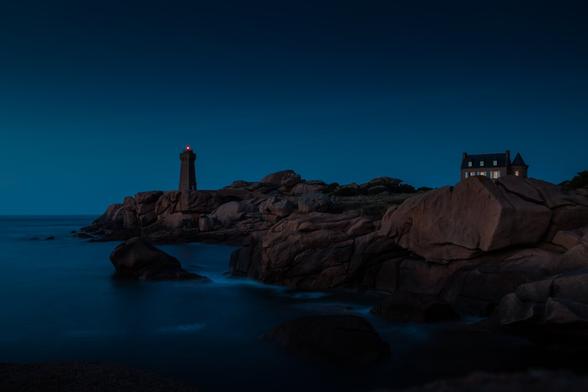 A lighthouse with a shining beacon on a rocky coastline at dusk, with a house nearby and calm ocean waters in the foreground.
