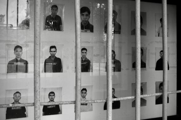 das schwarzweißbild im querformat zeigt durch gitterstäbe gesehen eine ausstellung von fotos von insassen des sog. s21-gefängnisses in pnomh penh.
an der brust tragen die personen schilder mit datum und häftlingsnummer.