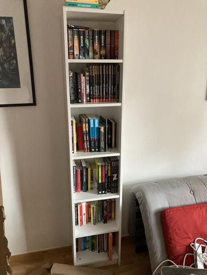 White , tall, thin bookcase packed with books / graphic novels.