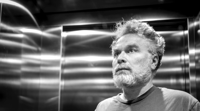 Monochrome photo of a bearded old man in a metal lift, the surface of which has streaks of reflected light