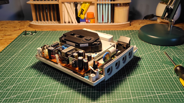 Zdjęcie przedstawiające konsolę SEGA Dreamcast ze zdjętą górną częścią obudowy przygotowana do serwisu zasilacza oraz nasmarowania mechanizmu napędu optycznego. Można też zauważyć, że przednia prawa podpórka obudowy pękła ze względu na stary wiek. Konsola stoi na zielonej macie antystatycznej.