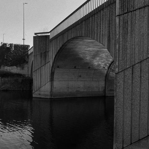 Photographie en noir et blanc, un pont avec une arcane où se glisse une rivière. 
La lumière reflète les mouvements de l'eau.