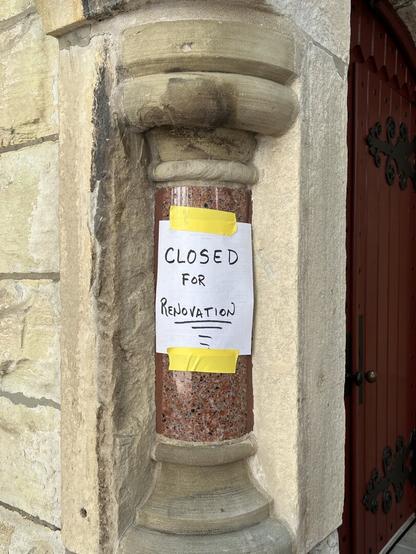 A red-pink marble column set in a stone corner of a chapel with red doors with black iron. On the column is a sign that says "Closed for Renovations."