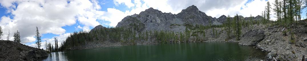 Panorama shot of a mountain Lake in Okanagan-Wenatchee National Forest.  A Green lakes stands in front of the viewer, surrounded to mostly by rocks and scattered trees. In the background is sharp mountains but on the left hand side you can see the reflection of the mostly cloudy sky in the water where it appears that there is nothing holding the water back