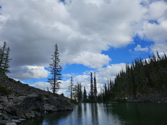 The reflection of trees can be seen in a slightly disturbed lake surrounded by sloping rocks.  The sky is mostly foggy but patches of blue peer through.