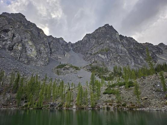 A lake is directly in front of a naked mountain ridge that goes up and down between various peaks. At their base our large piles of rocks and the occasional scattered tree