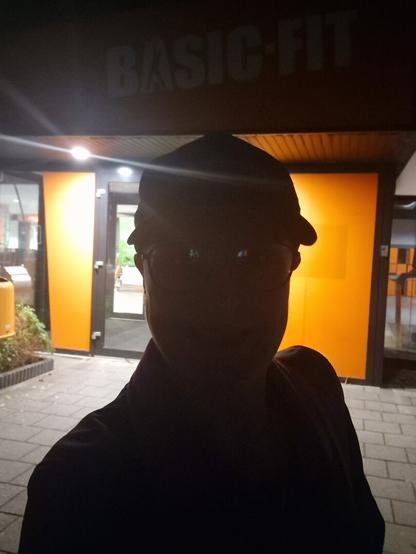 A person standing in front of the entrance to the Basic-Fit. A bright light in the back at the door, so their face seems darker. They're wearing a cap and glasses