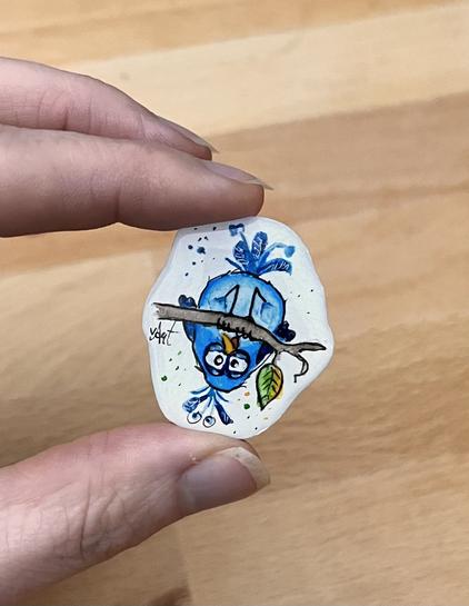 Eine Aquarellzeichnung mit Epoxidharz überzogen, circa 2 × 3 cm groß. Ich halte sie zwischen Damen und Zeigefinger. Ein blauer Vogel mit wirrem Blick, der Kopf über auf einem Ast sitzt. Seine Federn stehen ihm etwas wild vom Kopf ab