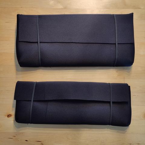 Two black envelope style sleeves for keyboards, with wrap around elastic closure, in black scuba fabric. On a wooden table