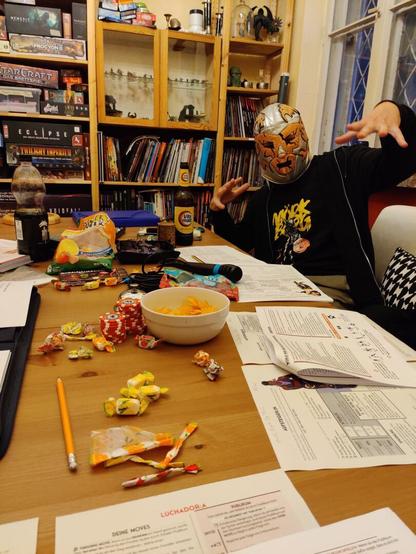 Tisch mit den typischen Rollenspiel Utensilien. Ein Spieler wirft sich mit Luchador Maske in Pose.