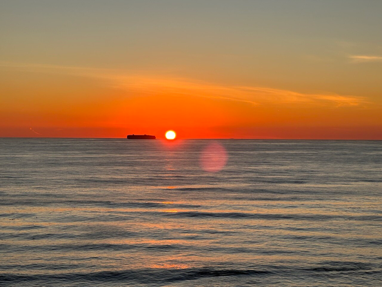 Blue water, bright orange sky, a boxy ship on the water just to left of orange setting sun