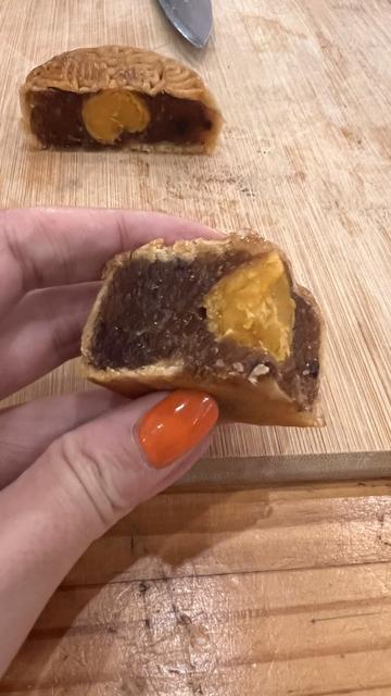 El pastelillo partido en dos. Una mano sostiene un cuarto al que le falta un mordisco. En el centro del pastelillo se ve una yema de huevo. Alrededor la jalea es color café. La mano es de piel blanca y tiene uñas naranja brillantes. Se ve una tabla de picar y un cuchillo.