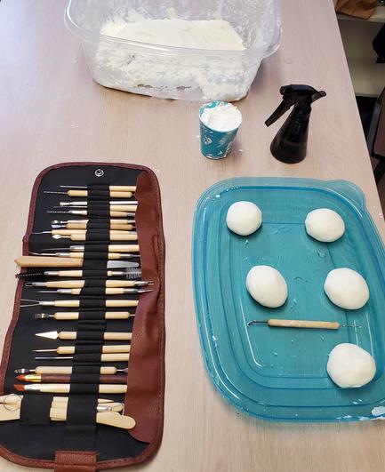 At bottom:
Sculpting tools in roll up pouch(on left).
Blue plastic lid with 5 white air dry clay balls & one sculpting hand tool(on right).
At top:
Plastic bin with white air dry clay.
Cup with white flour
Black water bottle with spray nozzle.