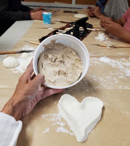 My left hand holding up a container of pre-made air dry clay - it is tan in colour. My DIY air dry clay is in front & shaped into a heart shaped tray.
In background are other people's hands working on different clay hand sculpting.