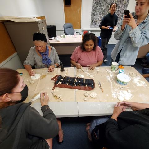 Overhead shot of women working on air dry clay creations at a table. Young facilitator on far right taking photo with her phone. Another young facilitator is in background.
