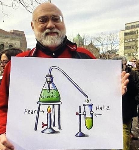 A man holding a poster with some chemistry equipment drawn upon it. A Bunsen burner is heating a flask labeled “Ignorance”. It is being evaporated by the burner, “Fear” and is condensed into a test tube labeled “Hate”.