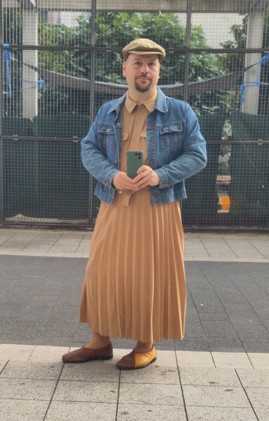 Selbstporträt in spiegelnder Scheibe. Mann mittleren Alters mit untersetzter Figur und dunklem Kinnbart. Ich trage ein beigefarbenes knöchellanges Kleid mit plissiertem Rockteil, ein blaue Jeansjacke, zweifarbig beige/braune Schuhe und eine hellbraune Mütze. Das Oberteil des Kleides ist wie ein Hemd geschnitten, mit Kragen, Knopfleiste und Brusttaschen. Die Schuhe sind schlicht zum Einschlupfen geschnitten, wie hoch geschlossene Ballerinas, mit dunkler Innenseite und hellerer Außenseite, mit vorne asymmetrischer Naht, die schräg nach außen läuft.