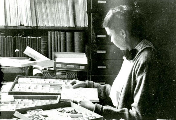 Historical black and white photo of a woman scientist working in her office. There are boxes with fossil specimens on her desk.