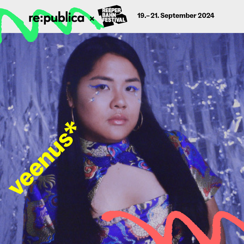 Das Foto ist ein Portrait von Vietnamesisch-Deutscher Sängerin und Produzentin veenus. Die Gesamtstimmung ist verträumt durch einen schimmernden, glitzernden Hintergrund in mattblau. In der Mitte des Bildes schaut veenus ernst in die Kamera. Sie hat lange schwarze Haare und ein vietnamesisch traditionell inspiriertes Oberteil aus Seide an. Die Seide ist mit goldenen Drachen bestickt, das typisch vietnamesische Design zeigt einen Halskragen.