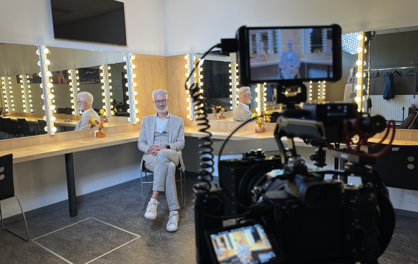 Me sitting in the dressing room for an interview at Semantics 2024 conference. I'm sitting in the corner of the room, mirrors of the dressing room on both visible walls. In the front (not exactly in the focus of the camery, but a bit blurry), we see the camera, which is used to record my interview with two small monitors, also showing me how I am sitting there. I am wearing a sand coloured 3 part suit, white shirt and white snearkers.