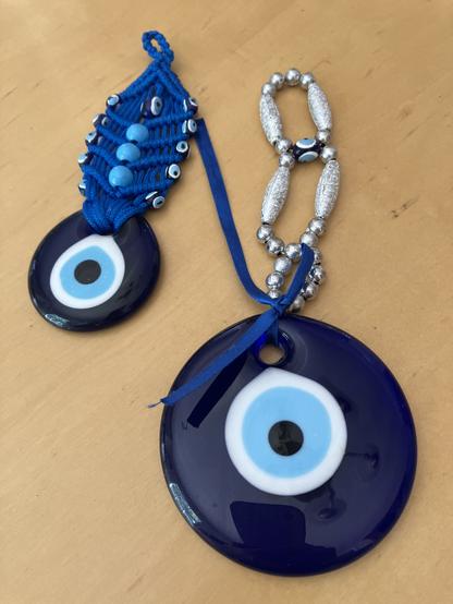 Two blue eye bead amulets (nazars) made from blown glass. The larger one hangs from a string of small silver beads, and the smaller one hangs from a blue macrame cord decorated with many small wooden eye beads.