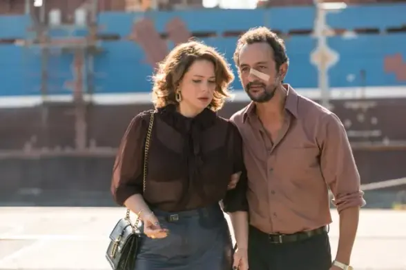 A man and a woman walk side by side. He has a bandage on his nose, and is gripping her arm. She is looking down