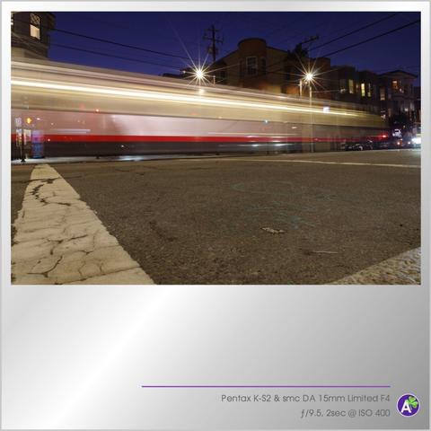 a color outdoor photograph. a train or subway streaks along a city street in the evening. this is a long exposure photograph, so the details of the passing train are blurred away. streetlights have the appearance of 14-point stars. there are no people or animals in this photograph.