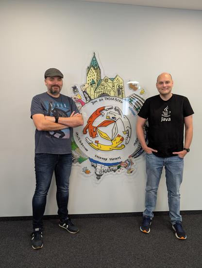 Zwei Personen stehen vor einer Wand. In der Mitte zwischen beiden Personen ein Kunstwerk