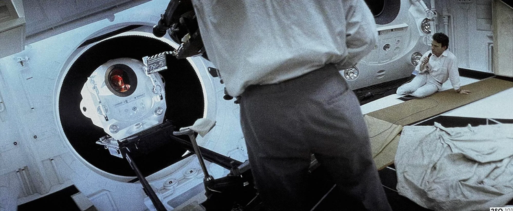 2001: A Space Odyssey (1968), as photographed by Kubrick. Dave (Keir Dullea) is in a small pod in the door of a white spaceship [no spoilers]. We're behind the cameraman, his camera is aimed at Keir. We can see a clapperboard and safely out off the shot a man in white – perfectly blending in with the ship – speaking with the actor via a handheld microphone. The shot in 28mm mimics the gorgeous 70mm widescreen format.
