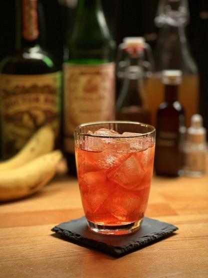 bright red cocktail with grated nutmeg garnish sitting on a black coaster in front of some bananas and several bottles: Doctor Bird pot still rum, Dolin rouge vermouth, banana-infused vermouth, coconut fat-washed Campari, tepache de piña and a small dropper of saline