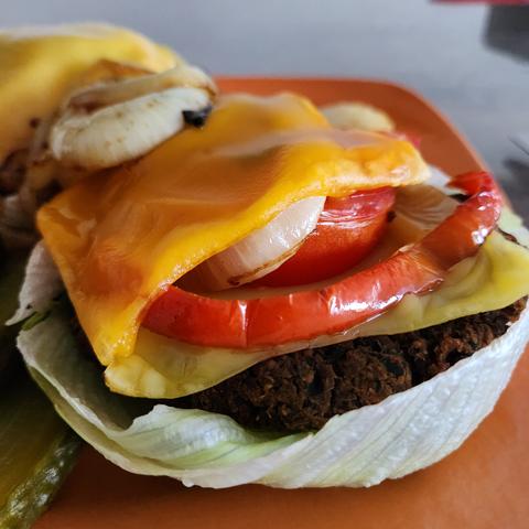 Rote-Bete-Burger auf einen organefarbenen Teller angerichtet. Garnitur Gewürzgurken,  gebratene Zwiebeln und roter, gebratener Paprika. 1 Burger auf einem Salatblatt gebettet, Nahaufnahme und mit veganen Käse überbacken. Glutenfrei und vegan.